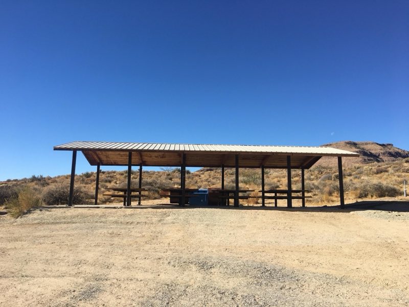 A shade structure consisting of beams holding up a solid roof. Underneath are six picnic tables.