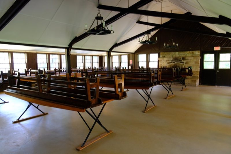 Wooden tables and benches inside a building with a stone fireplace and numerous windows