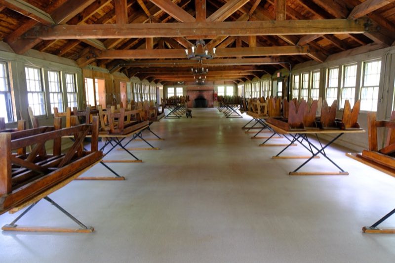 Inside of a large wooden cabin with two lines of benches and tables and a brick fireplace at the end