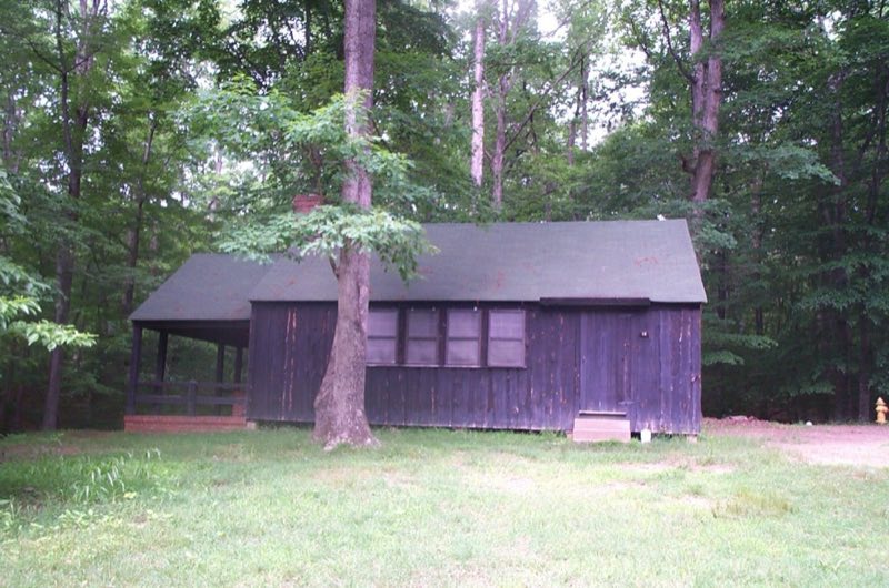 Wooden cabin building with a brick porch and chimney shaded by a green forest