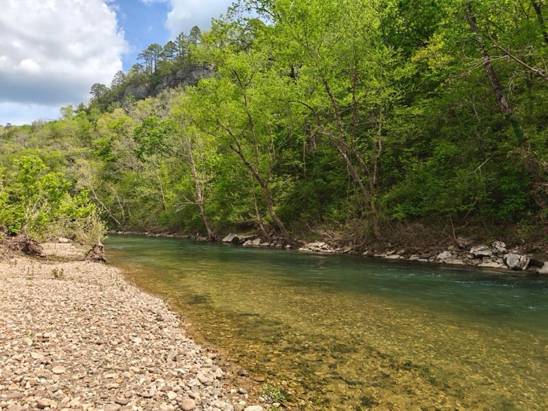 The image shows a shallow, clear river flowing through a rocky area, with a wide pebble-covered bank