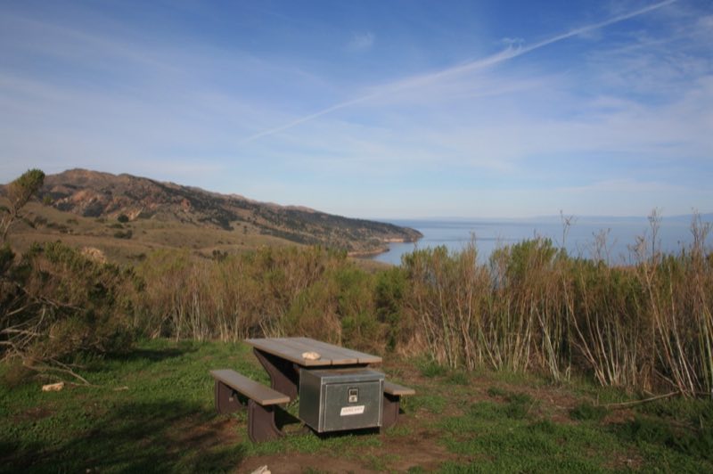 Campground picnic table on green grass overlooking ocean, coastline, and blue sky with white clouds