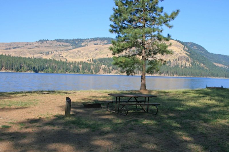 picnic table and fire pit next to campsite number post, with lake in background