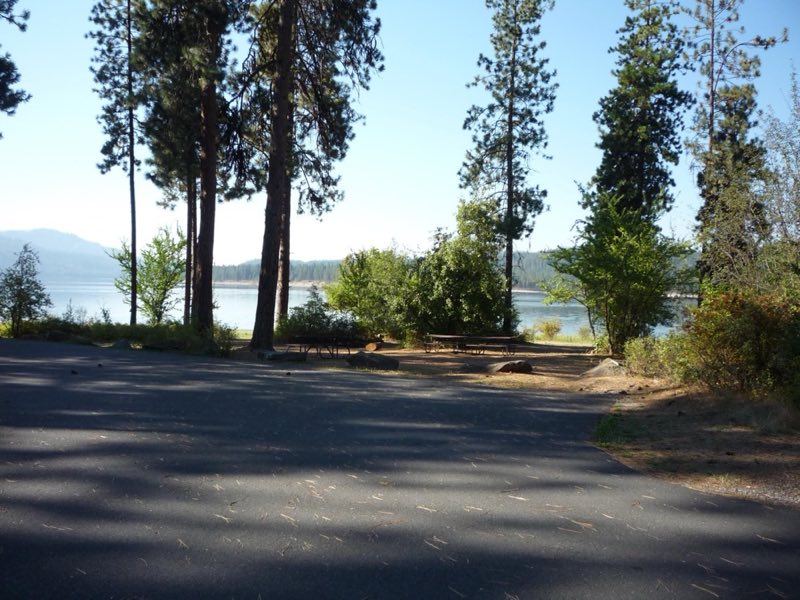 A campsite with a paved parking area, picnic table, fire ring, and lake access.