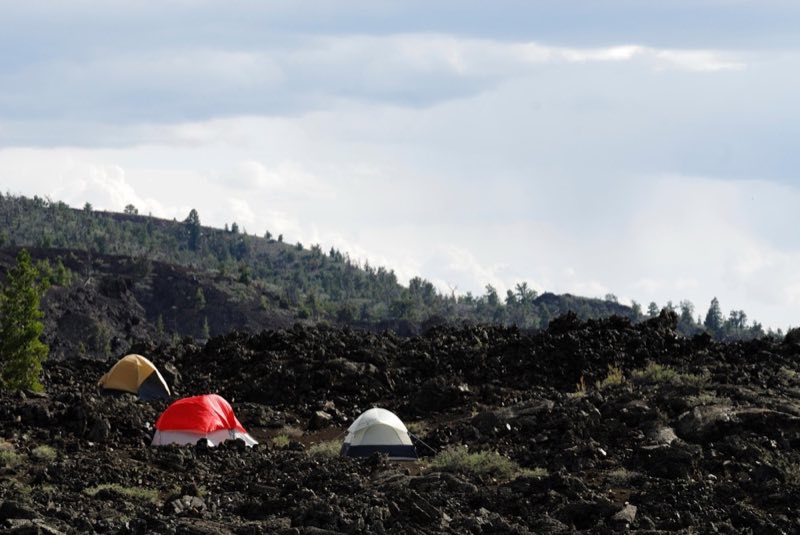 three tents on a hill of black lava rocks with a dark hill covered with trees in the distance