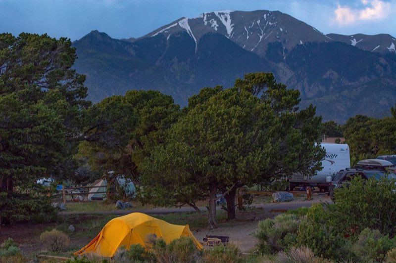 A tent and RV in a campground with small trees, with a snow-capped mountain in background