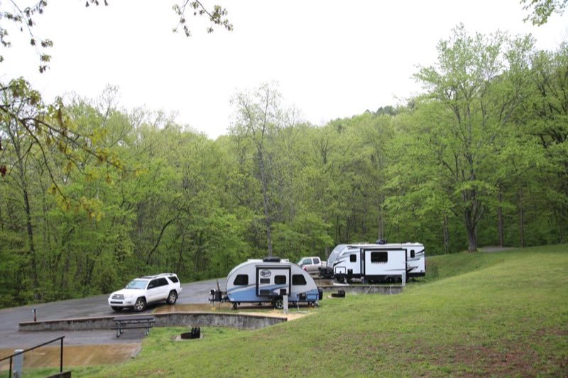 Two campers/RVs sit on concrete pads. A large forest surrounds the campsites.
