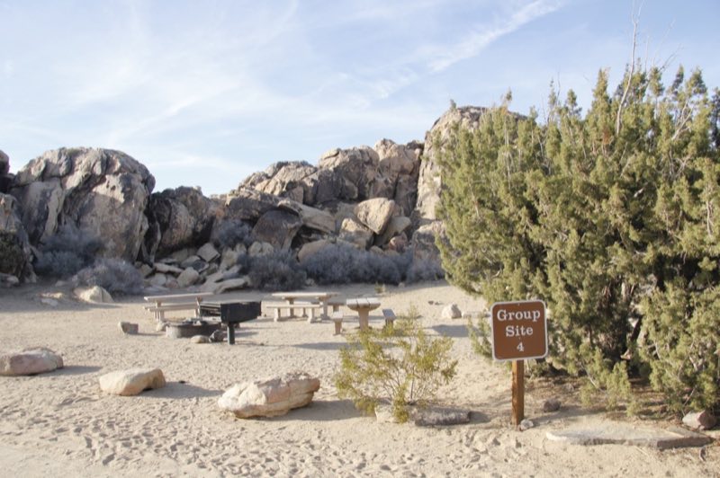 A sign that reads "Group Site 4". Behind are three picnic tables, a fire ring and a fire grate.