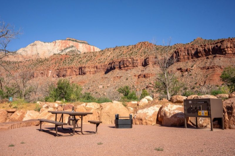 A gravel campsite lined with boulders. A picnic table, fire ring, and storage box sit in the site.