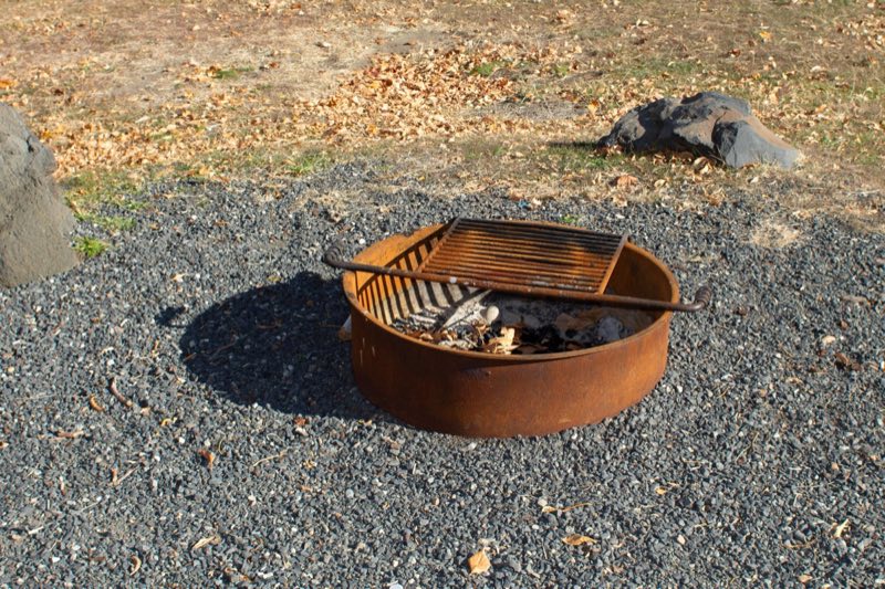 Round rusted metal ring on the ground with a slated grate over the top used for grilling and fires.