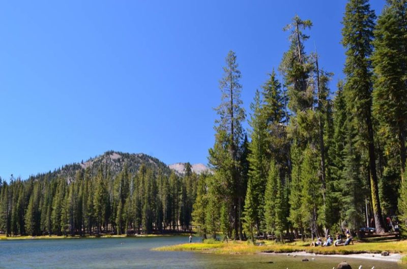 People sit and stand on a grassy shore of a mountain lake.