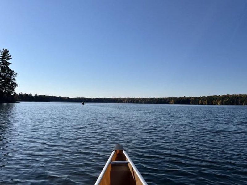 The front of a canoe surrounded by a blue lake with another canoe in the distance.