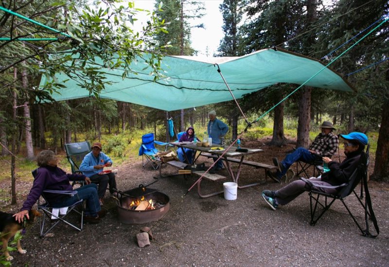 several people in camp chairs around a fire, under a tarp hung from trees