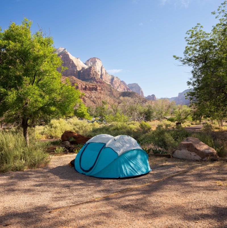 A blue tent sits in a gravel campsite. In the background, huge sandstone cliffs are visible.