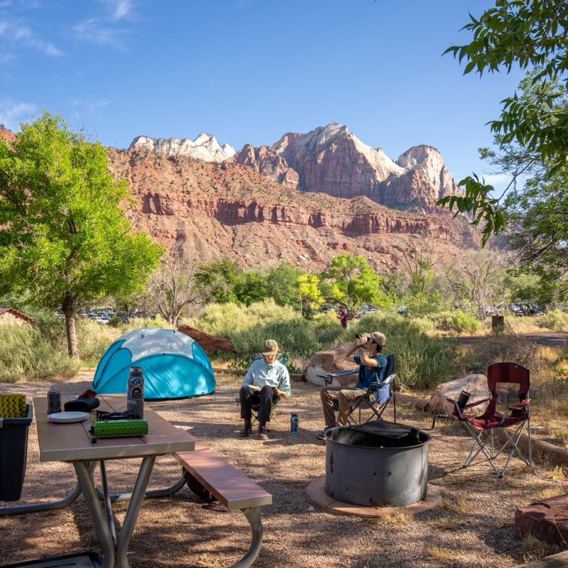 A man and woman sit in a campsite. In the background are multicolored sandstone cliffs.