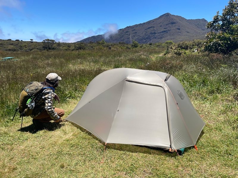 camper setting up tent in grassy area with mountain peak in background